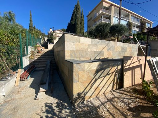 Mur de pierre et escalier menant à un bâtiment dans un cadre résidentiel. Ciel bleu.