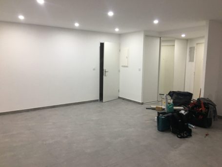 Espace vide avec murs blancs, sol gris, porte noire et matériel de construction.
