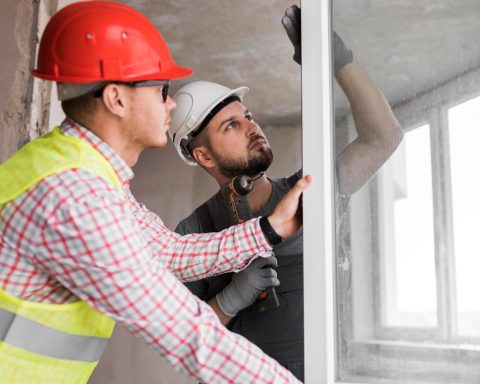 Deux ouvriers en chantier posent une vitre, portant casques et gilets de sécurité.