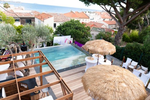 Piscine avec chaises longues et parasols, vue sur des maisons et la mer.